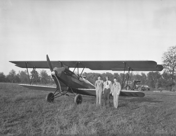Black-and-white photograph of a biplane parked on a grassy field at Halley Field, with three men standing beside the aircraft.